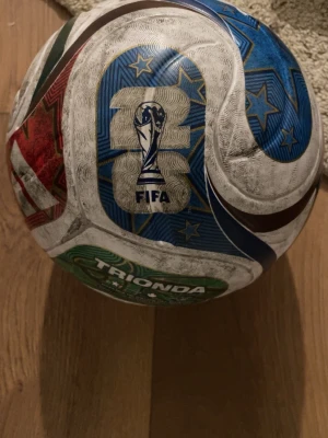 FIFA fotboll Trionda blå/vit/röd - Säljer en fotboll med FIFA-logga och färgglada mönster i blått, rött, grönt och vitt. Bollen har stjärndetaljer och texten 'TRIONDA' på en av panelerna. Perfekt för fotbollsspel på gräs eller konstgräs.   Kan tvätta den så den ser helt ny ut innan den säljs pris kan diskuteras. . Storleken är 5