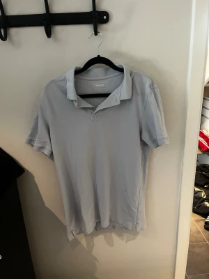 Ljusblå pikétröja från Dressmann - En enkel och stilren ljusblå pikétröja från Dressmann med klassisk krage och korta ärmar. Tillverkad i mjuk bomull som känns skön mot huden. Perfekt för en avslappnad och clean look.