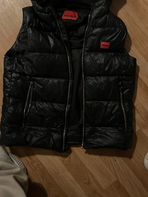 Svart dunväst från Hugo - Snygg svart dunväst från Hugo med röda logodetaljer på bröstet och insidan. Västen har två dragkedjeförsedda fickor och hög krage. Perfekt för lager på lager och ger en clean streetwear-look.
