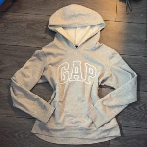 Grå hoodie GAP - Snygg grå hoodie från GAP med stor vit broderad logga på bröstet. Mjuk insida och klassisk huva. Perfekt för en avslappnad och trendig stil. Passar till jeans eller mjukisbyxor. Skriv om ni har några frågor och priset går att diskuteras