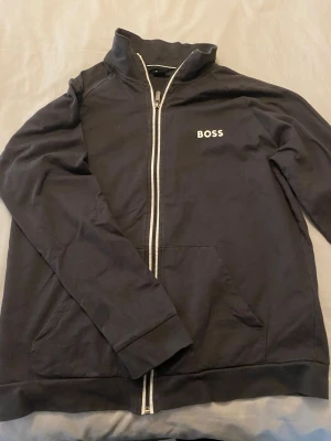 Svart zip-hoodie från BOSS - Svart hoodie från BOSS med vit dragkedja och logga på bröstet. Klassisk modell med fickor framtill och ribbade muddar. Perfekt för en clean och stilren look. Materialet känns mjukt och bekvämt.