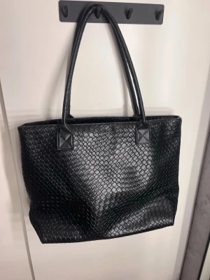 Svart shoppingväska i flätad look - Stilren svart shoppingväska med flätad struktur och dubbla axelband. Väskan har en rymlig rektangulär form och är tillverkad i konstläder som ger en lyxig känsla. Perfekt för dig som vill ha plats för allt du behöver under dagen.