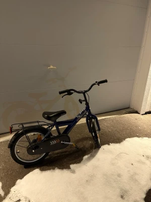 Blå barncykel med pakethållare - Säljer en blå barncykel med svart sadel och styre. Cykeln har pakethållare bak, kedjeskydd med pilmönster och reflexer på hjulen. Passar perfekt för yngre barn som vill cykla runt i området.