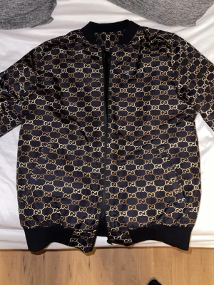Svart bomberjacka  - Snygg svart bomberjacka med guldfärgat GG-mönster över hela jackan. Jackan har ribbade muddar, dragkedja framtill och klassisk bomberjacke-passform. Materialet har en glansig finish som ger en exklusiv känsla.