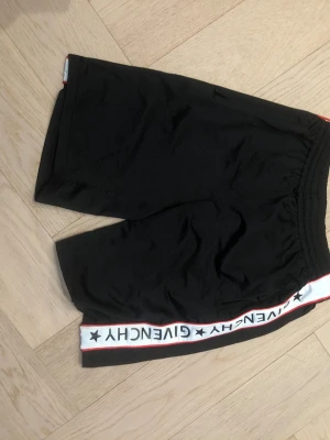 Svarta shorts från Givenchy - Säljer ett par svarta shorts från Givenchy med vit och röd sidoremsa där det står GIVENCHY och stjärnor. Shortsen har elastisk midja och sportig look, perfekta för en avslappnad stil.