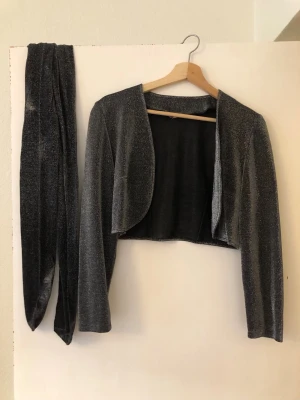 Glittrig bolerotopp i mörkgrått - Säljer en glittrig mörkgrå bolerotopp med lång ärm och matchande bälte/skärp. Kort och öppen framtill, perfekt för att lyfta en outfit med lite extra shimmer. Materialet är mjukt och stretchigt syntet med metallic-effekt. Med i köpet även ett par matchande glittriga strumpbyxor storlek L
