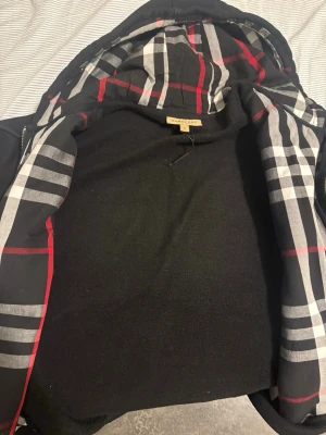 Svart zip-hoodie från Burberry - Säljer en svart hoodie med dragkedja från Burberry. Insidan av huvan har det klassiska rutmönstret i svart, vitt och rött. 100% äkta, bästa kvaliten. Pickup tc eller post. Tvek inte att ställa frågor