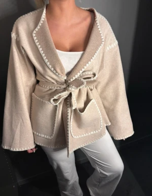 Beige kappa AWARE - Snygg beige omlottjacka från Aware med knytbälte i midjan och dekorativa vita sömmar längs kanterna. Jackan har två stora fickor framtill och en avslappnad, loose passform. Perfekt nu till hösten och vintern. Nypris 900kr, mycket fint skick, pris går att diskutera🤍