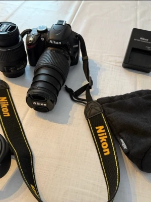 Nikon D3200 systemkamera med två objektiv - Nikon systemkamera D3200 med två utbytbara objektiv i svart utförande. Kamerahus med skärm på baksidan och standard Nikon-rem. Ett zoomobjektiv med brännvidd 18–55 mm och ett telezoomobjektiv med längre brännvidd 55-200mm båda med Nikon F-fattning. Medföljer gör även objektivlock, motljusskydd, kamerarem, laddare och mjuk förvaringspåse till objektiv eller tillbehör. Utrustningen lämpar sig för allmän fotografering, till exempel vardagsbilder, resor och enklare natur- eller porträttfoto.  Jag fi