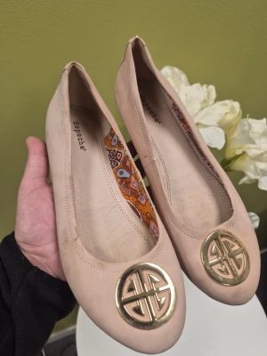 Depeche rosa  ballerinaskor med gold detaljer 38 - Snygga beige ballerinaskor från Depeche med rund tå och dekorativt guldfärgat metallmärke framtill. Skorna har ett färgglatt paisleymönstrat innerfoder som ger en unik touch. Tillverkade i ett mjukt material som känns bekvämt på foten.
