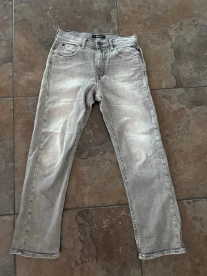 Grå raka jeans från Replay - Säljer ett par gråa jeans från Replay med raka ben och klassisk femficksmodell. Jeansen har slitna detaljer och ljus tvätt för en avslappnad look. Materialet är denim. Dem är 70cm i midjan och 90cm lång.
