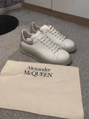 Vita Alexander McQueen sneakers - Stilrena vita sneakers från Alexander McQueen med tjock sula och grå detalj vid hälen. Skorna har klassisk låg profil, perforerade sidor och är tillverkade i slätt läder. Perfekta för dig som gillar exklusiv och modern streetstyle.