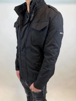 Svart vindjacka från Woolrich - Svart vindjacka från Woolrich med hög krage, flera fickor framtill och axelklaffar. Jackan har lång ärm och stängs med dragkedja och knappar. Märkespatch på ärmen och ribbade muddar vid ärmslut och nederkant. Perfekt för dig som gillar en stilren och funktionell look.