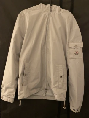 Ljusgrå vindjacka från Moncler - Snygg ljusgrå vindjacka från Moncler med huva. Jackan är i jättebra skick, den helt felfri och är använd cirka 2-3 gånger. Nfc och qr kod funkar som det ska. Storlek M, passar bättre som S. Hör av er vid frågor och funderingar😀