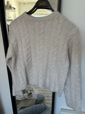 Ljusbeige kabelstickad tröja - Använd ca 2 gånger tidigare. Mysig kabelstickad tröja i ljusbeige med rund hals och breda ärmar. Tröjan har ett klassiskt flätmönster framtill och på ärmarna, vilket ger en trendig och avslappnad look. Perfekt att matcha med jeans för en chill vardagsstil. 