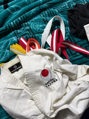 Karatedräkt med bälten Budoworld - Säljer en vit karatedräkt från Budoworld med japansk flagga och broderad text på jackan. Ingår flera bälten i vitt, gult, orange, rött och rödvitt. Dräkten är i klassisk modell med byxa och jacka i kraftigt bomullstyg, perfekt för träning eller cosplay.