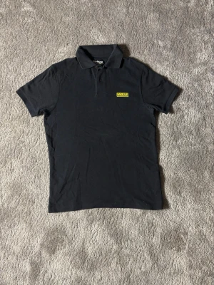 Svart pikétröja från Barbour S - Klassisk svart pikétröja från Barbour International med gul logotyp på bröstet. Tröjan har krage, knappar vid halsen och korta ärmar. Perfekt för en clean och stilren look.