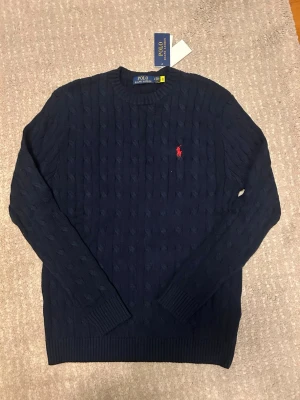 Mörkblå kabelstickad tröja Polo Ralph Lauren - Mörkblå kabelstickad tröja från Polo Ralph Lauren med rund halsringning och röd broderad logga på bröstet. Tröjan har långa ärmar och ribbade muddar vid ärmslut och nederkant. Perfekt för en stilren och klassisk look.