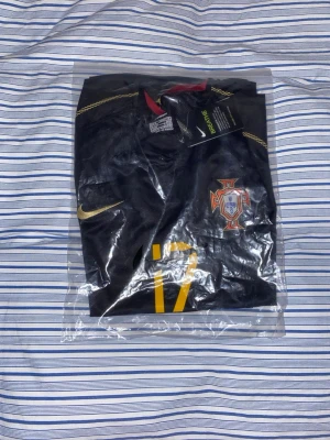 Portugal svart matchtröja Nike #17 - Säljer en svart Portugal matchtröja från Nike med nummer 17 i gult på framsidan. Tröjan har gulddetaljer, Portugal-emblem på bröstet och officiell turneringspatch på ärmen. Tillverkad i lätt och ventilerande material, perfekt för fotbollsträning.