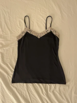 Svart linnetopp med spetsdetalj - Stilren svart linnetopp med tunna axelband och dekorativ beige spets längs urringningen. Toppen har en v-ringad framtill och ärmlös design som ger ett elegant och feminint uttryck. Den är i fint skick men kommer tyvärr inte till användning hos mig, och jag hoppas därför att den kan komma till glädje hos någon annan. Priset är inte hugget i sten, så hör gärna av dig vid intresse eller frågor.