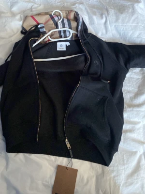 Svart Burberry hoodie med dragkedja - Snygg svart hoodie från Burberry med dragkedja och klassiskt rutigt foder i huvan. Hoodien har en broderad Burberry-logga på bröstet och en stor ficka framtill. Tillverkad i mjukt bomullsmaterial och har guldfärgade detaljer vid dragkedjan.