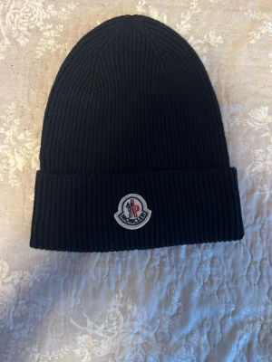 Mörkblå ribbstickad mössa från Moncler - Mörkblå ribbstickad mössa från Moncler med uppvikt kant och klassisk Moncler-logga framtill. Mössan är tillverkad i mjukt material och har en enkel, stilren design som passar perfekt till streetwear och vardagsstil. Som nyskick, endast testad.