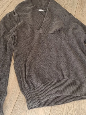 Grå stickad tröja från Bläck - Mysig grå stickad tröja från Bläck med bred sjalkrage och ribbade muddar. Perfekt för lager på lager och har en avslappnad passform. Tröjan är i ett mjukt material och passar till många olika stilar.