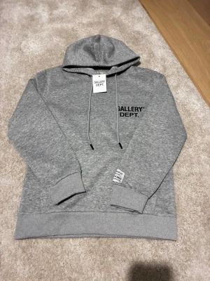 Grå hoodie från Gallery Dept. - Snygg grå hoodie från Gallery Dept. med svart logga tryckt på bröstet. Klassisk modell med huva och dragsnören samt ribbade muddar vid ärmar och nederkant. Tillverkad i mjukt material som känns skönt mot huden. 