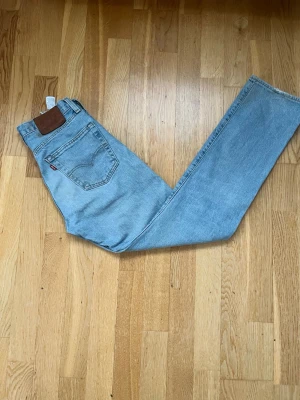 Ljusblå raka Levi's jeans - Säljer ett par klassiska ljusblå Levi's jeans med rak passform och hög midja. Jeansen har fem fickor, Levi's ikoniska läderpatch bak och röd logotagg på bakfickan. Tillverkade i slitstark denim, perfekta för en avslappnad och trendig look.