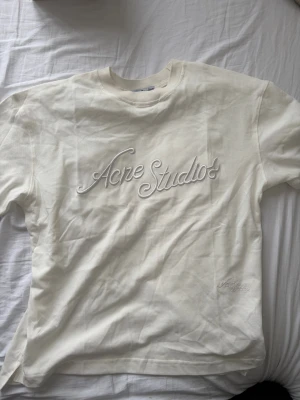 Vit t-shirt från Acne Studios - Säljer en stilren vit t-shirt från Acne Studios med broderad logga framtill. T-shirten har rund halsringning och korta ärmar. Tillverkad i mjuk bomull för en skön känsla och clean look. Perfekt för dig som gillar minimalistisk design.