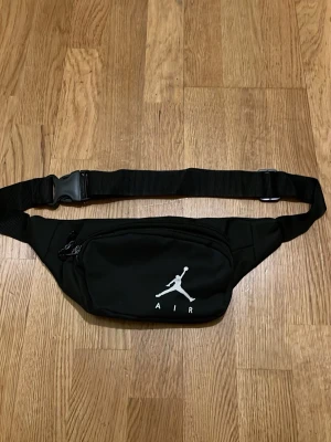 Svart midjeväska från Jordan - Snygg svart midjeväska från Jordan med klassiska Jumpman-loggan i vitt framtill. Väskan har justerbart band, två dragkedjefack och är tillverkad i slitstarkt tygmaterial. Perfekt för att bära det viktigaste när du är på språng.