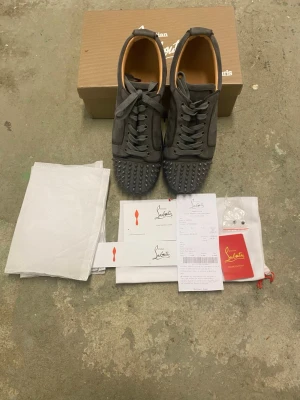 Gråa Christian Louboutin sneakers - Säljer ett par gråa sneakers från Christian Louboutin med ikoniska röda sulor och coola nitar på tån. Skorna har snörning och är tillverkade i mocka med läderdetaljer. Perfekta för dig som vill sticka ut med en lyxig och edgy stil.