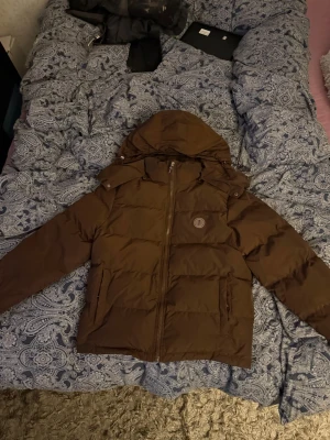 Brun pufferjacka från Trapstar - Säljer en brun pufferjacka från Trapstar med stor huva och broderad logga på bröstet samt Trapstar-text i ryggen. Jackan har dragkedja framtill, två sidofickor och är tillverkad i ett glansigt, vadderat material som håller värmen under kalla dagar.                   Säljer pågrund av att den är för liten för mig använd 2-3 gånger 