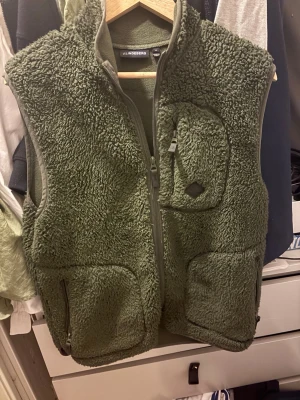 Grön fleeceväst från J.Lindeberg - Mysig grön fleeceväst från J.Lindeberg med hög krage och dragkedja framtill. Västen har tre fickor med dragkedja, varav en på bröstet. Perfekt för lager-på-lager och riktigt skön att bära under kyliga dagar.