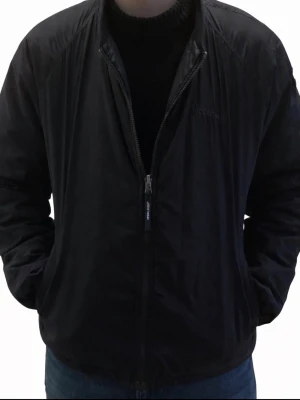 Svart vindjacka från Woolrich - Snygg svart vindjacka från Woolrich med diskret rutmönster och dragkedja framtill. Jackan har en klassisk rak passform, två sidofickor och broderad logga på bröstet. Perfekt för blåsiga dagar och enkel att matcha med olika outfits.