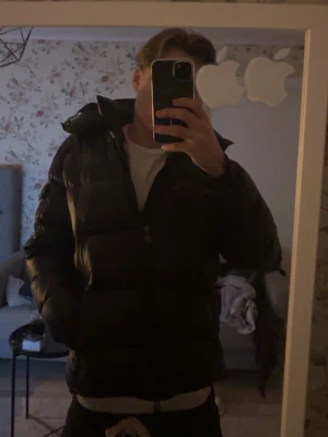 Moncler Maya - Säljer en svart pufferjacka från Moncler med klassisk quiltad design och huva. Jackan har Moncler-logga på ärmen och dragkedja framtill. Enda defekten är ett litet litet hål på vänstra sida bröst som är lagat med laglapp men det går enkelt att sy.   Storlek 2 S/M