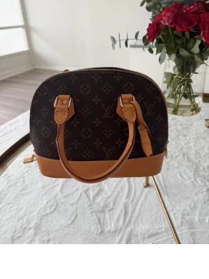 Handväska  - Säljer handväska i klassiskt brunt monogramcanvas med ljusbruna läderdetaljer och guldfärgade metalldelar. Väskan har två handtag, rundad form och dragkedja upptill. Perfekt för dig som gillar lyxig och tidlös design.