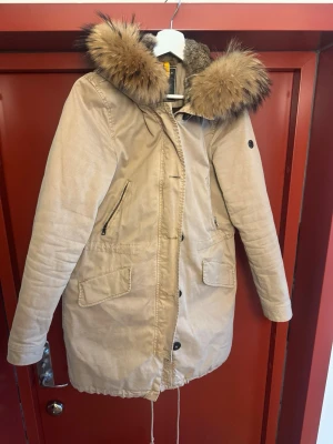 Beige parkas med pälsluva från BLONDE no. 8 - Stilren beige parkas med stor huva kantad av fluffig päls. Jackan har dragkedja och knappar framtill, två stora fickor med lock och två snedställda bröstfickor med dragkedja. Justerbar midja och nederkant för perfekt passform. Perfekt för kalla dagar.