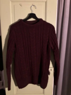 Vinröd kabelstickad tröja - Mysig vinröd kabelstickad tröja med rund hals och långa ärmar. Tröjan har ett klassiskt mönster och ribbade muddar vid ärmslut och nederkant. Perfekt för kalla dagar och enkel att matcha med jeans eller kjol.