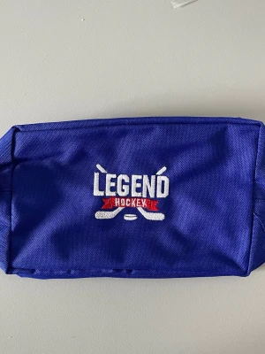 Blå necessär Legend Hockey - Blå necessär från Legend Hockey med broderad vit och röd logga framtill. Tillverkad i slitstarkt tyg och har en rektangulär form, perfekt för att förvara tejp eller andra hockey accessoarer. Snygg och praktisk för hockeyintresserade.