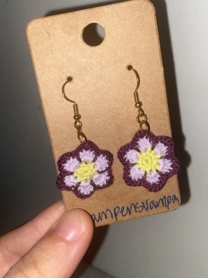 Handgjorda virkade blomörhängen - Unika örhängen med virkade blommor i lila, vitt och gult. Blommorna har fem kronblad och är fästa på guldfärgade krokar. Perfekta för dig som vill ha en färgglad och personlig accessoar.