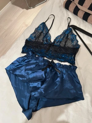 Blått spets- och satinsovset - Snyggt tvådelat set med blå spets-bh och matchande blå satinshorts. Bh:n har tunna axelband och vacker spetsdetalj i svart och blått. Aldrig använd 