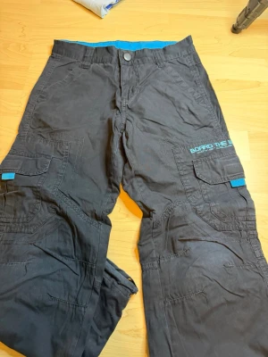 Svarta täckbyxor Board The World - Täckbyxor i svart med blå detaljer och tryck från Board The World. Byxorna har flera fickor med lock, justerbar midja och coolt grafiskt tryck på baksidan. Perfekta för vintersport och streetstyle.