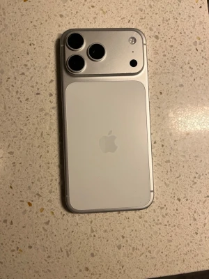 Vit iPhone med tredubbel kamera - Snygg vit iPhone med stilren design och rundade hörn. Baksidan är i glas med Apple-loggan och har ett kamerahus med tre linser och blixt. Metallramen ger en exklusiv känsla och framsidan har en stor skärm som täcker nästan hela ytan. Perfekt för dig som gillar modern teknik.