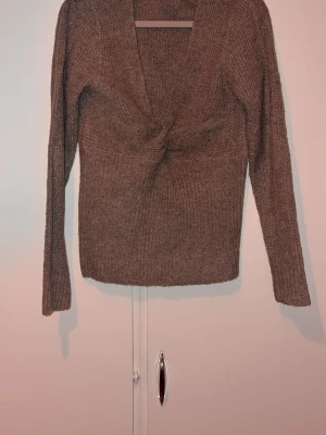 Brun stickad tröja med twist framtill - Säljer en brun stickad tröja med lång ärm och snygg twistdetalj framtill. Tröjan har djup v-ringning och är ribbstickad för en skön passform. Perfekt för dig som gillar en enkel men trendig look.