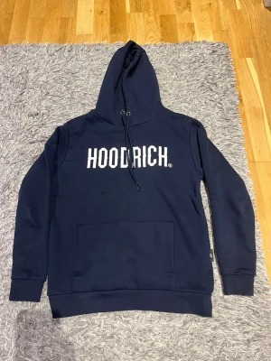 Marinblå HOODRICH hoodie - Snygg marinblå hoodie från HOODRICH med stor vit logga på bröstet. Klassisk känguruficka framtill, justerbar huva med snören och ribbade muddar vid ärmar och nederkant. Tillverkad i mjukt bomullsmaterial för maximal komfort.