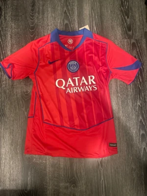 Röd PSG matchtröja Nike - Säljer en röd Paris Saint-Germain matchtröja från Nike med blå detaljer och Qatar Airways-tryck på bröstet. Tröjan har korta ärmar, rund hals och är tillverkad i ett lätt, ventilerande material perfekt för fotboll eller träning.