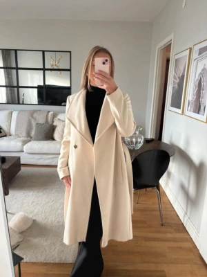 vit kappa med skärp - Snygg krämvit kappa med breda slag och knytbart midjebälte. Kappan har en rak, elegant passform och stängs med en dold knapp framtill. Den är i storlek M men passar S/xs