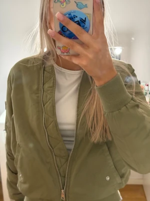 Olivgrön bomberjacka från Cropp - Snygg olivgrön bomberjacka från Cropp med klassisk ribbad mudd vid ärmslut och nederkant. Jackan har dragkedja framtill, två fickor med tryckknapp och quiltade detaljer. Perfekt för dig som vill ha en stilren och trendig look.