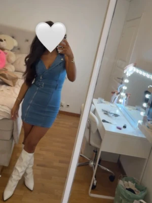 Blå jeansklänning med bälte - Snygg blå jeansklänning med v-ringning och bälte i midjan. Klänningen är kort och har breda axelband, perfekt för en trendig look. Passformen är tight och materialet är klassiskt jeans. Matcha med boots för en cool outfit.